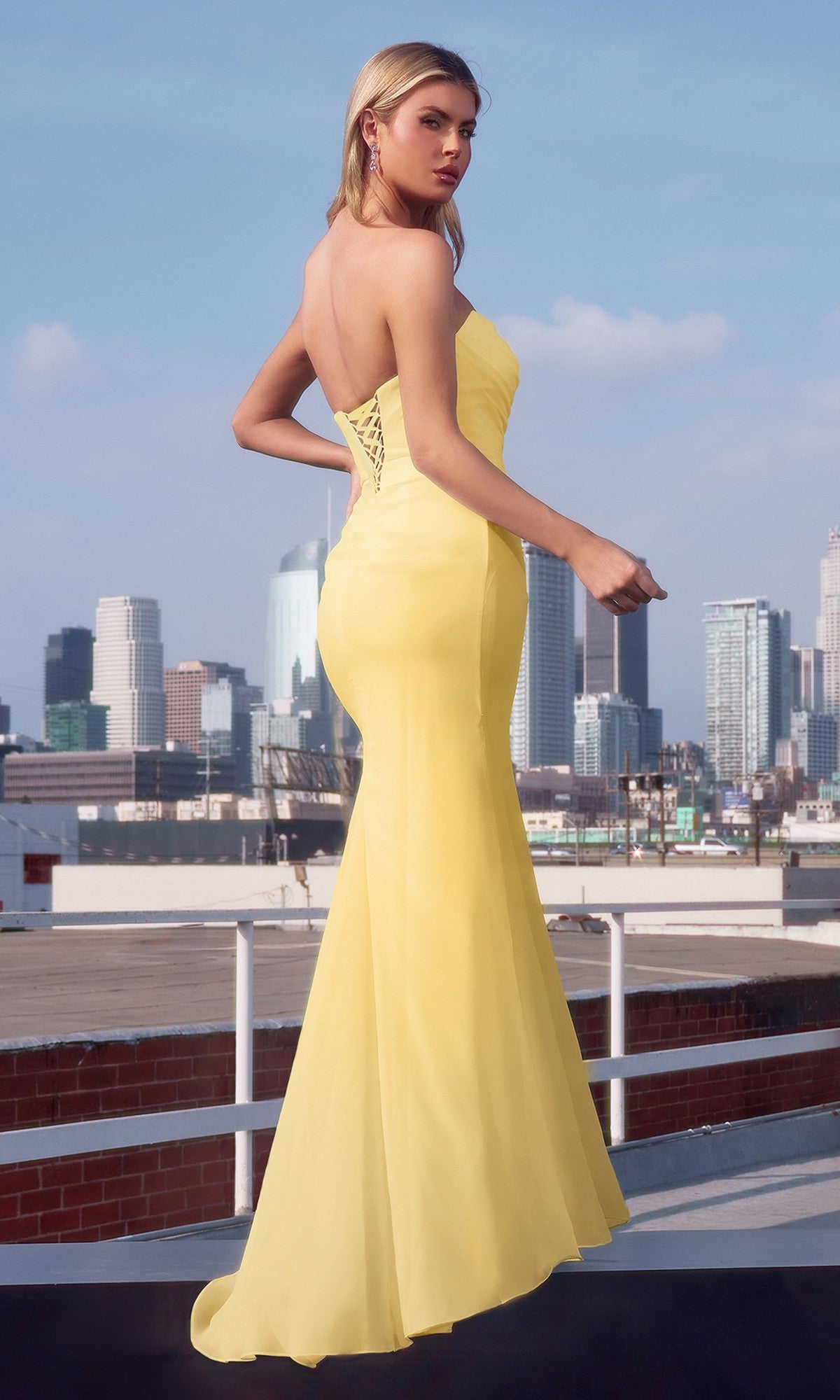 Wearing the Amoris T702W long prom dress, a woman stands on a rooftop in a flowing yellow chiffon gown, the strapless style and floor-length silhouette set against the city skyline.